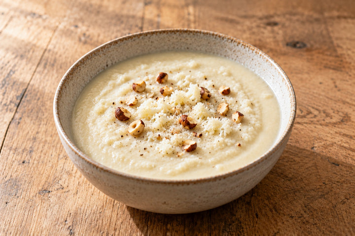 Velouté de chou-fleur blanc et crémeux dans un bol, parsemé de noisettes grillées concassées et de quelques sommités crues râpées